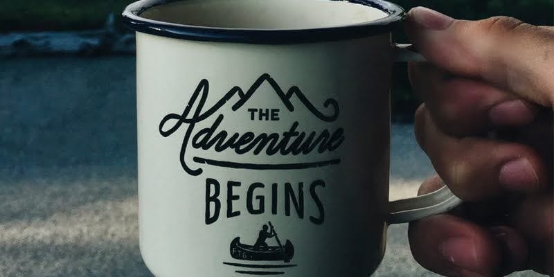 Hand holding a mug with 'THE ADVENTURE BEGINS' and mountain graphics, outdoors.