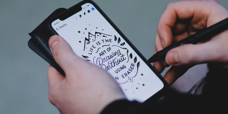 Person creating hand-lettered design on phone screen that reads 'Life is the art of drawing without an eraser.'